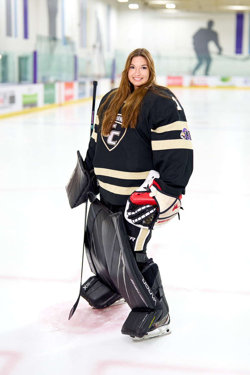 A smiling hockey goalie kneels on the ice wearing a dark jersey and full goalie equipment.