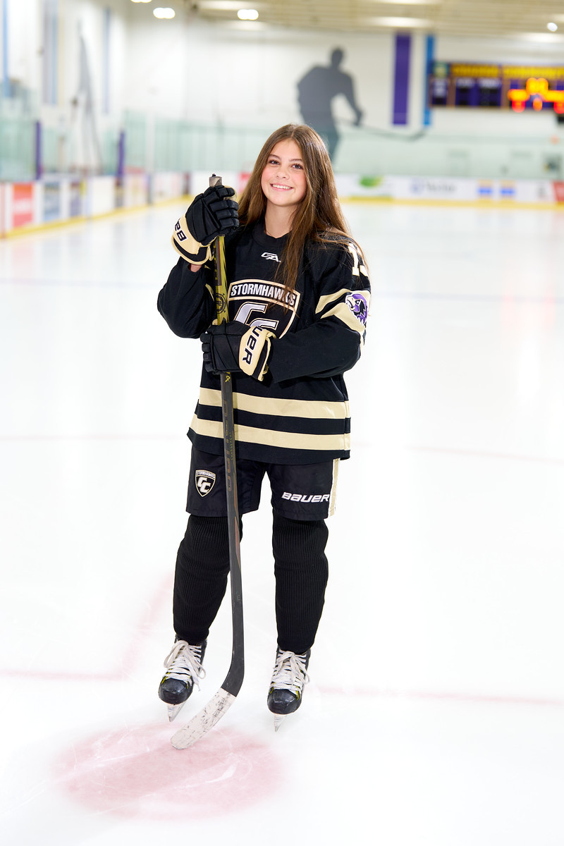 A smiling hockey player stands on the ice in a dark jersey with her hockey stick.