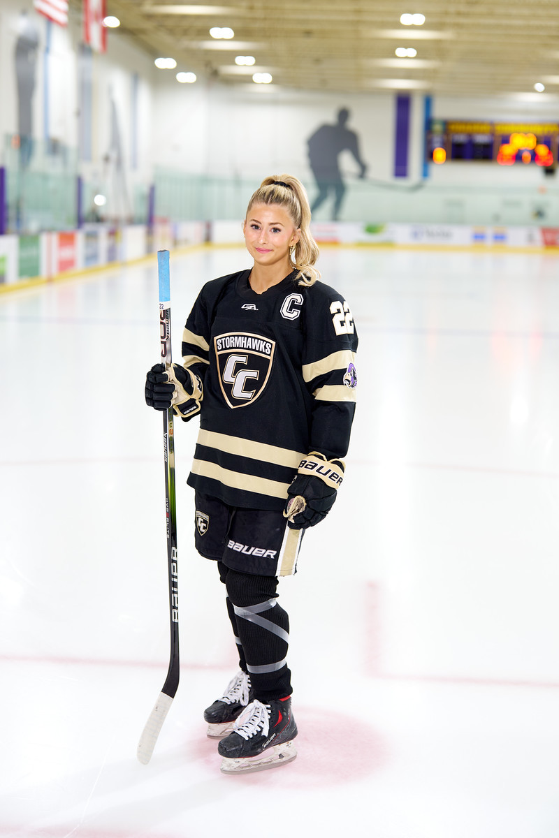 A hockey player poses on the ice in a black and gold uniform, holding her stick upright.