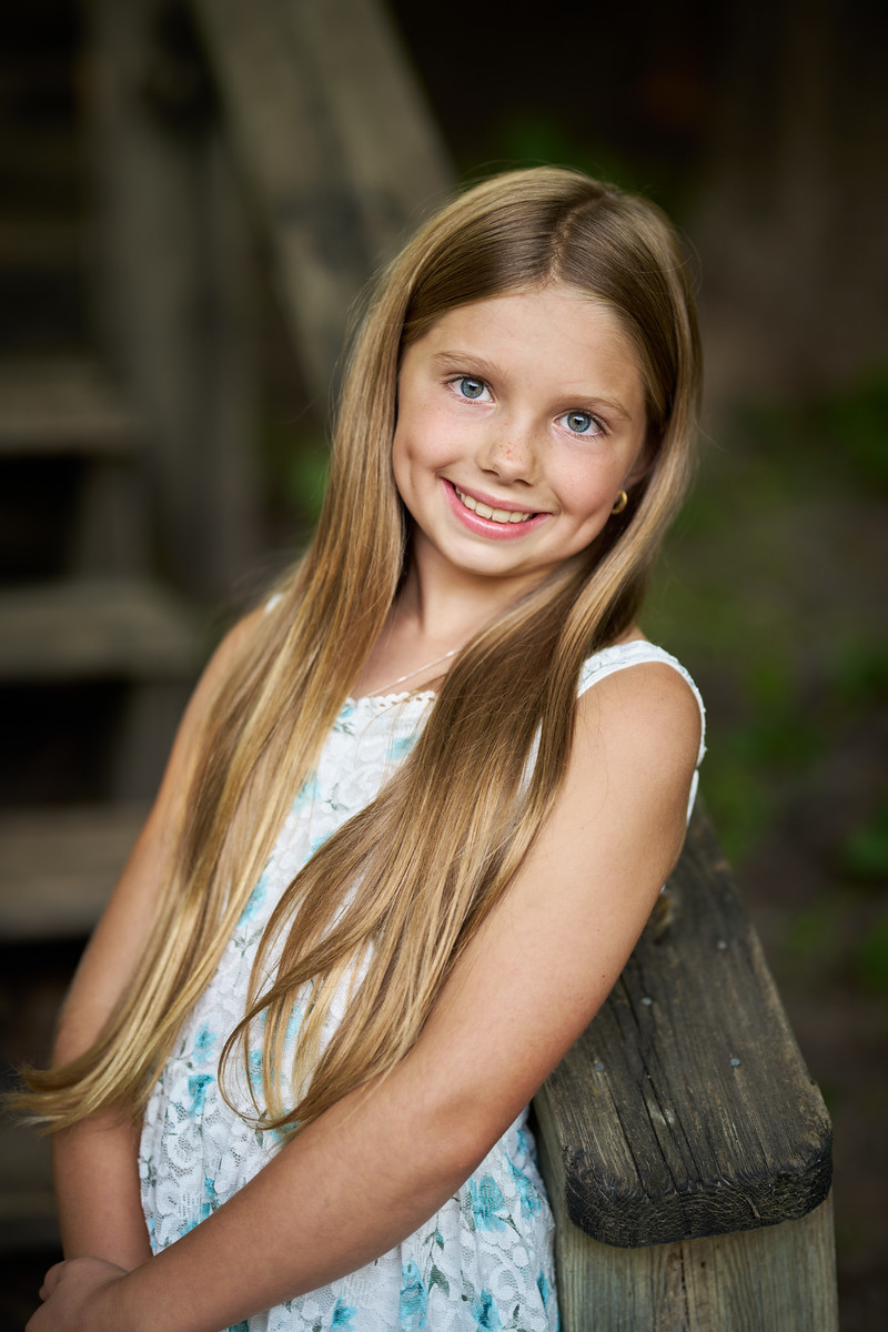 A smiling girl with long hair leans against a wooden post beside shaded steps.
