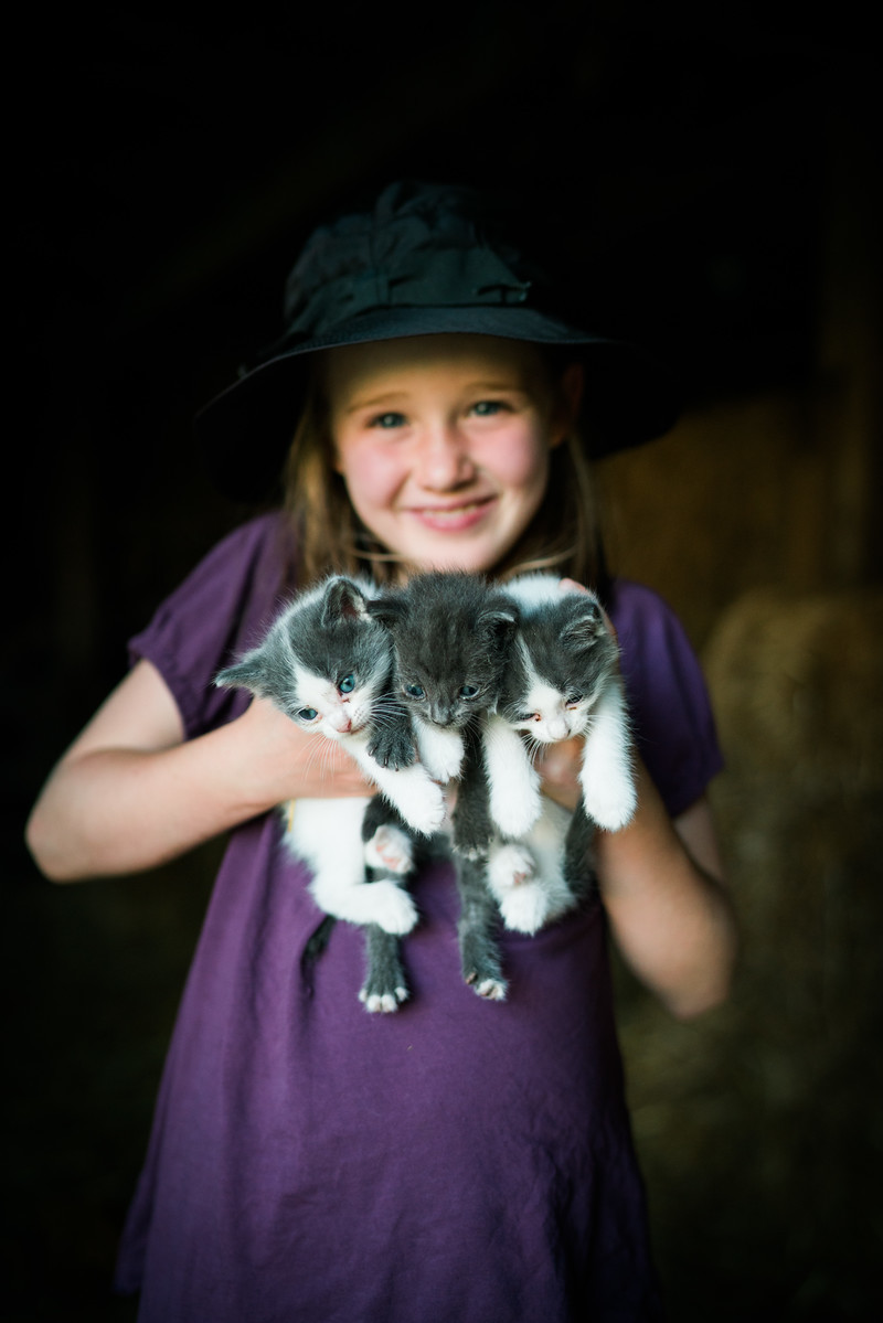 A smiling girl wearing a dark hat holds two small kittens against a shaded barn background.