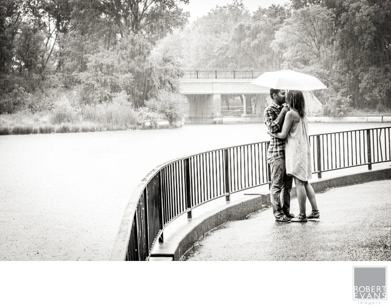 A quiet black-and-white portrait of a couple embracing beneath an umbrella, using the rain and surrounding landscape to create a timeless, intimate photograph.