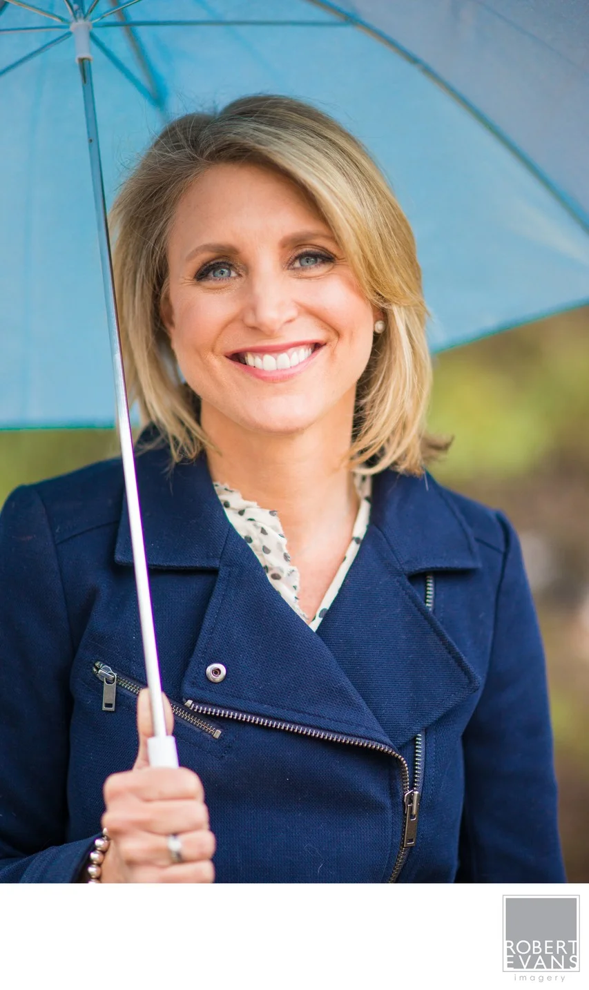 A bright, natural portrait of Belinda Jensen KARE 11, using a blue umbrella as both a visual element and frame, creating a relaxed photograph filled with personality.