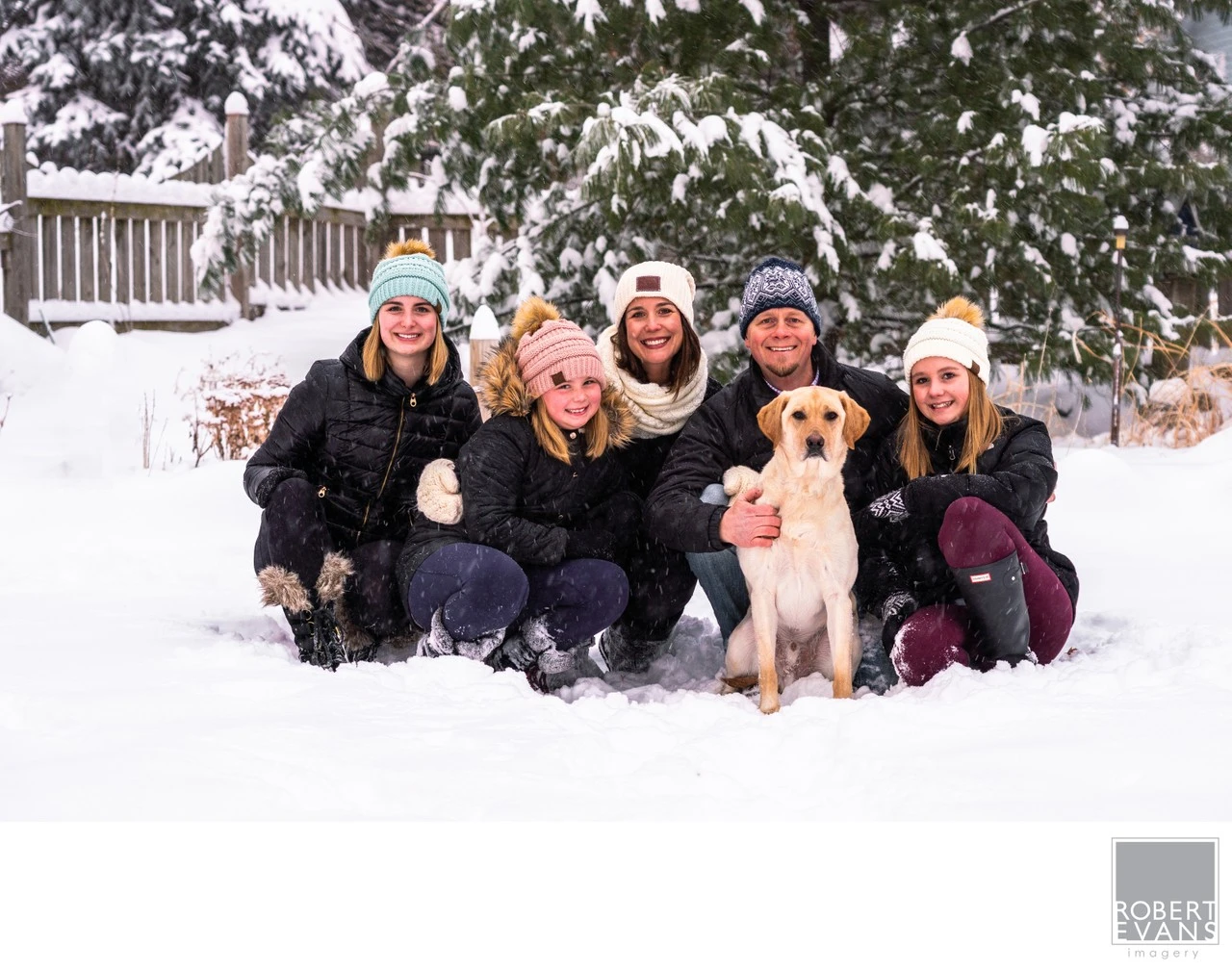 Parents and three daughters sit together in fresh snow with their dog between them.