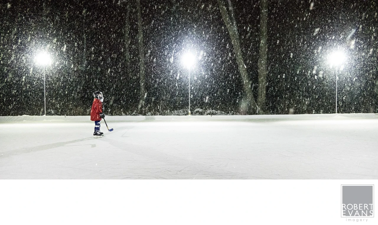 A young hockey player stands alone on an outdoor rink during a heavy Minnesota snowfall, creating a dramatic winter portrait that captures childhood, sport and life in Minnesota.
