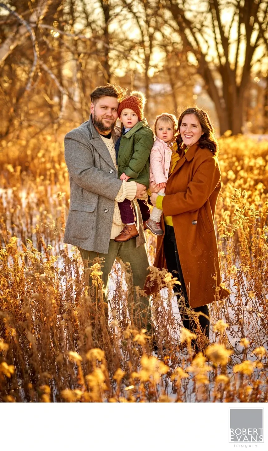 Parents and two young children stand together among sunlit autumn trees and tall plants.