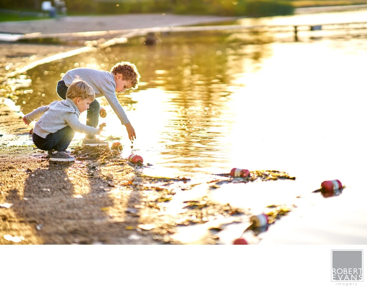 A young boy bends toward shallow water beside a sandy shoreline in warm evening light.