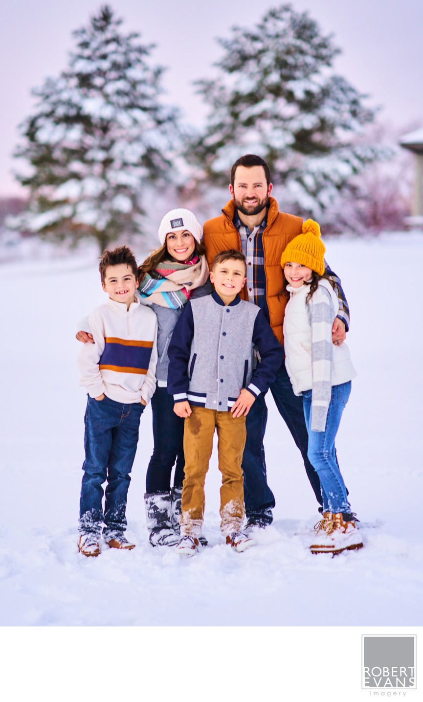 A relaxed winter family portrait featuring parents and their three children together in the snow, surrounded by snow-covered Minnesota evergreens.