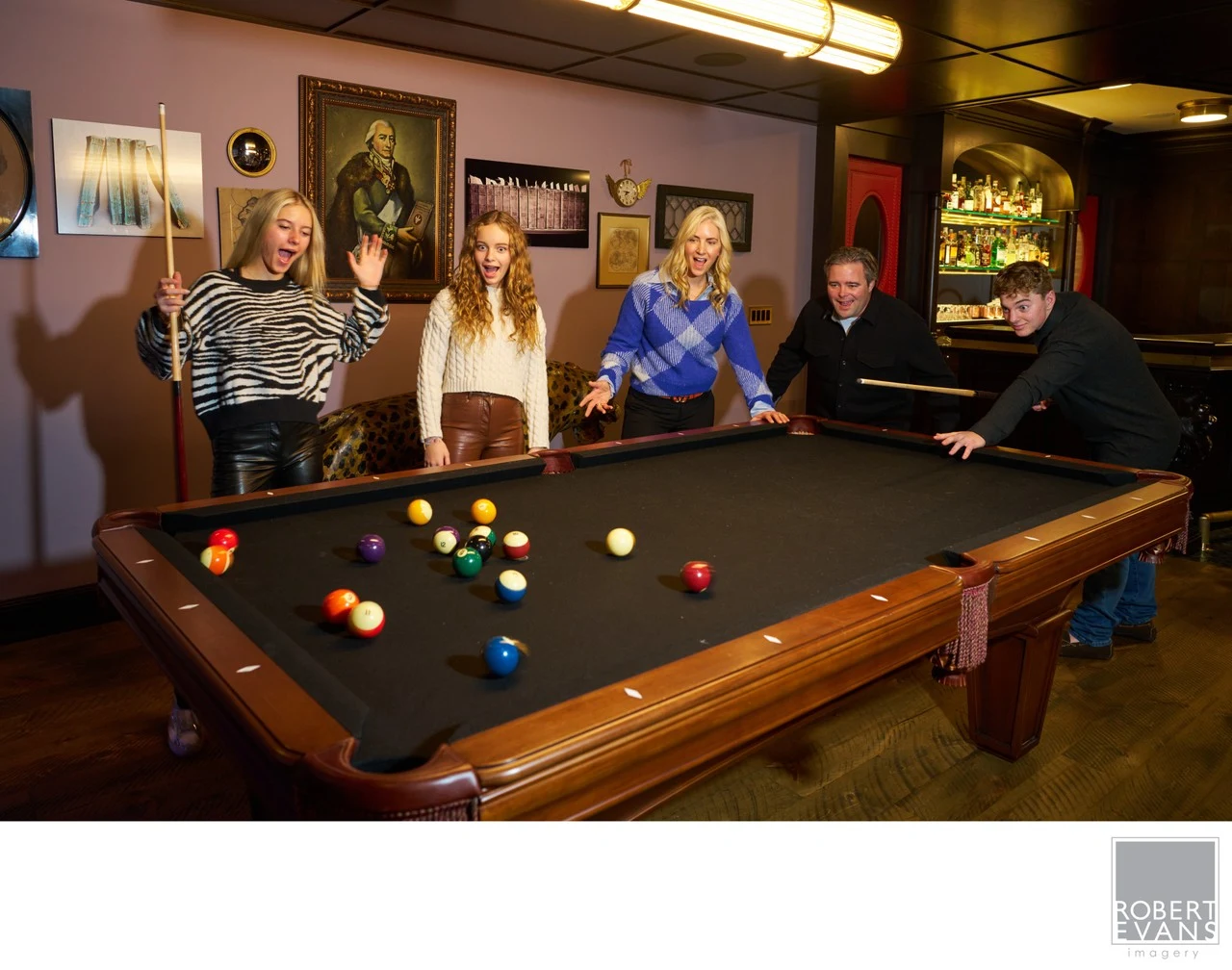 Five family members gather around a pool table during a relaxed portrait at home.
