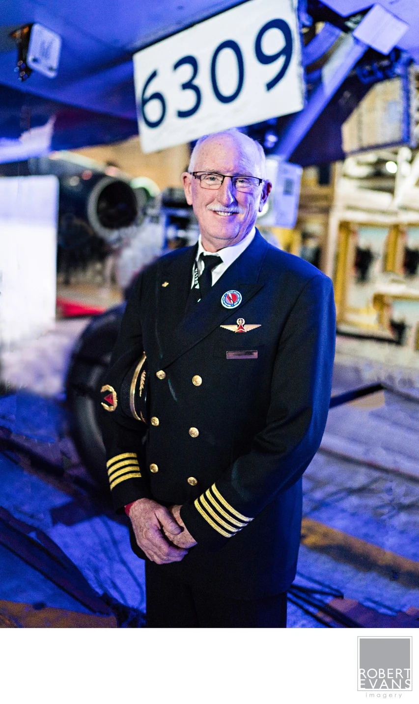 An older man in a pilot uniform poses beside an aircraft inside a hangar.
