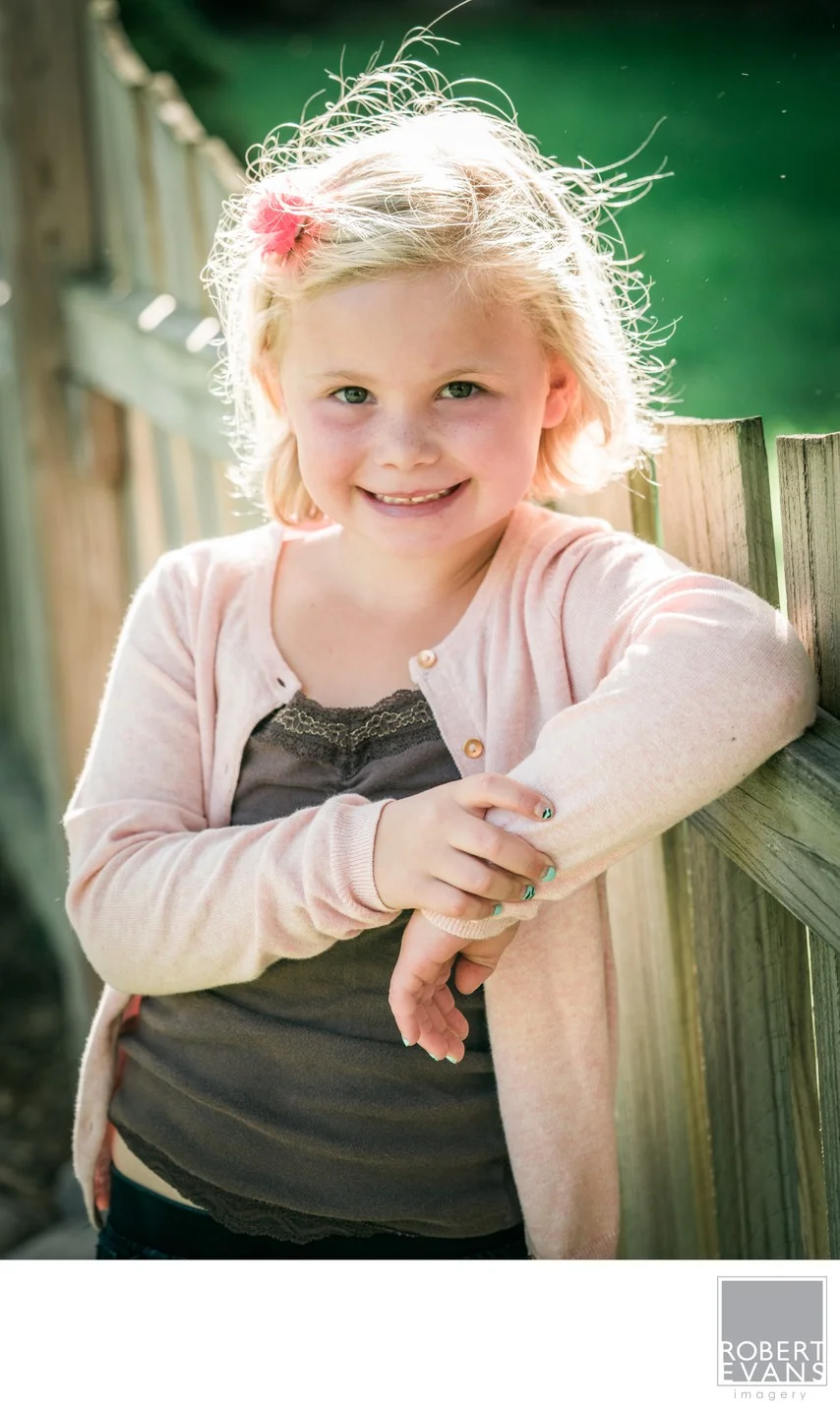 A smiling little girl in a pale pink cardigan leans against a wooden railing.