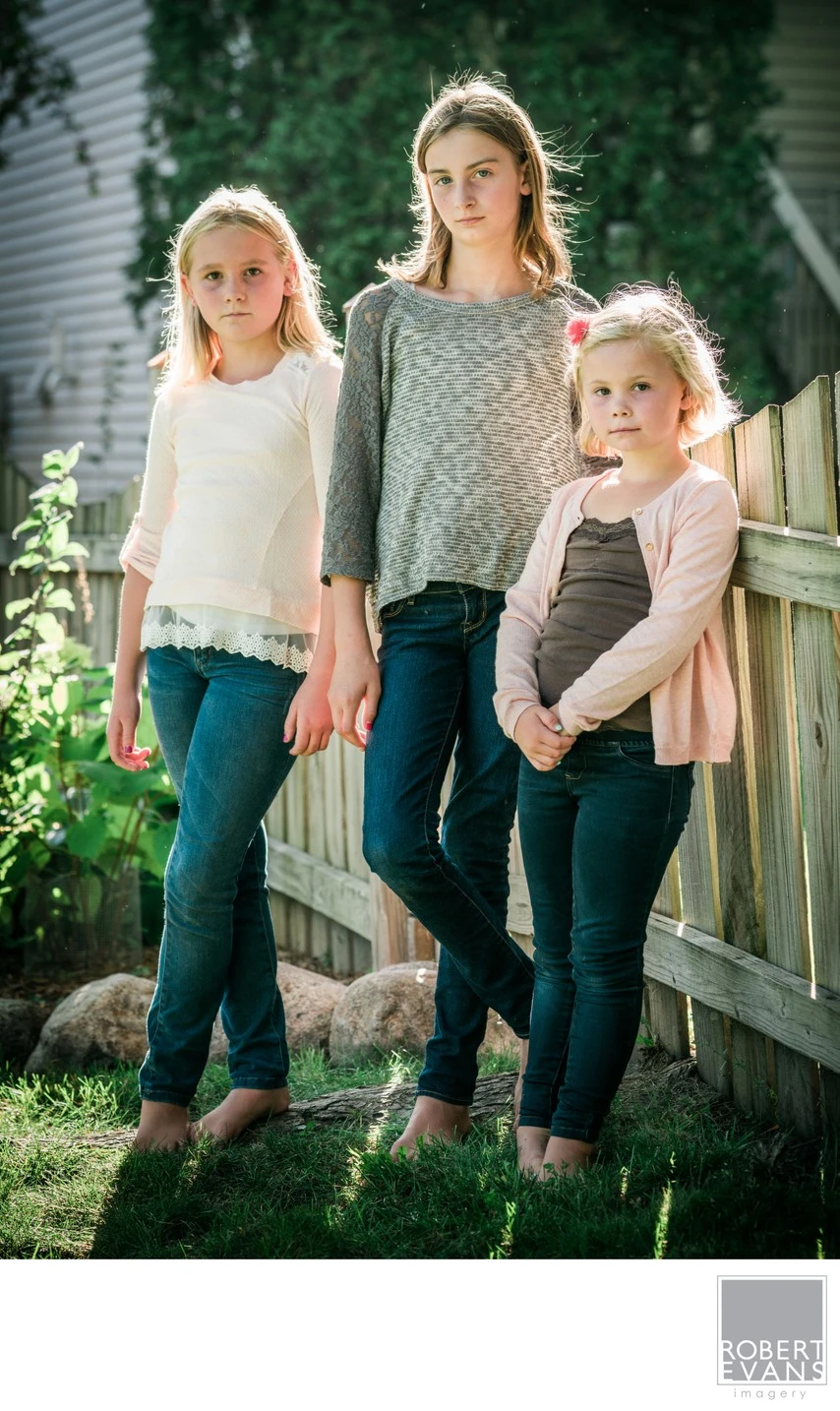 Three girls stand together beside a wooden porch railing in soft outdoor light.