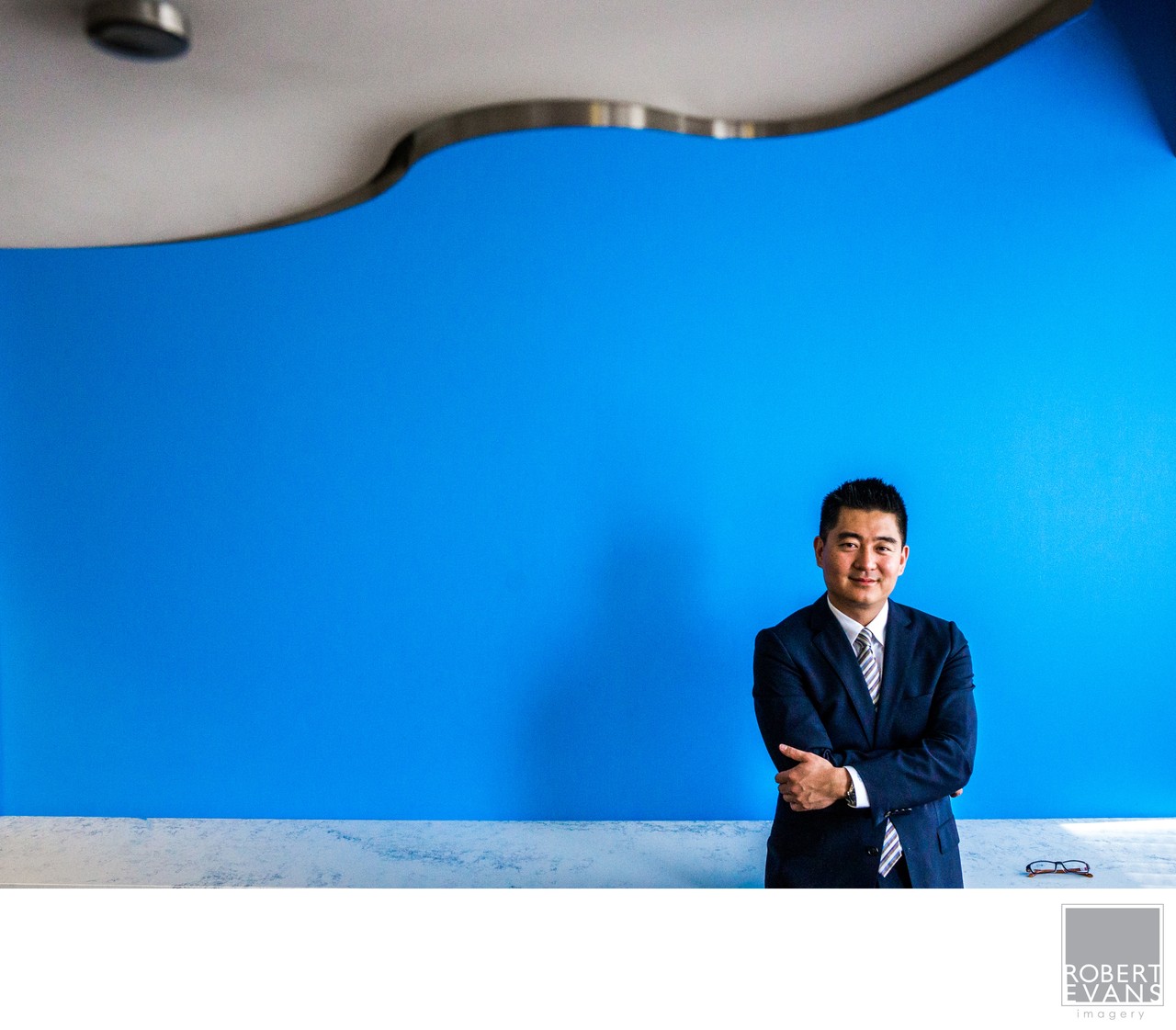 A man in a dark suit stands against a vivid blue wall beneath a curved architectural detail.