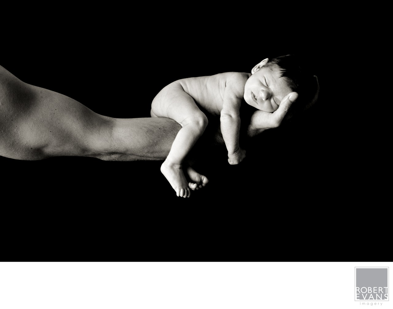 A tiny newborn sleeps across an adult’s outstretched hand against a dark background.