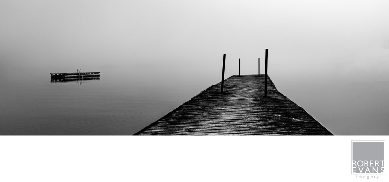 A wooden pier extends into a misty lake in black and white.