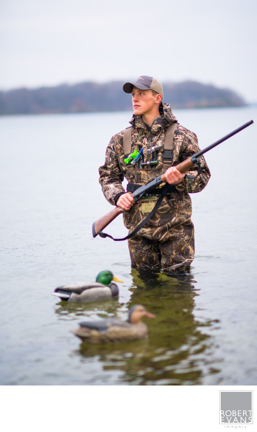 A hunter in camouflage stands in water with a shotgun and duck decoys.