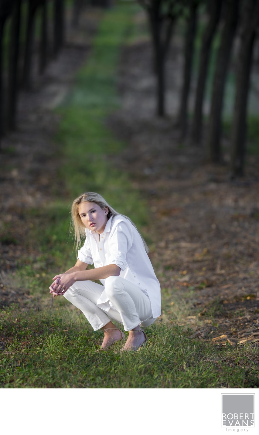 A woman dressed in white crouches beside a path beneath a long canopy of trees.