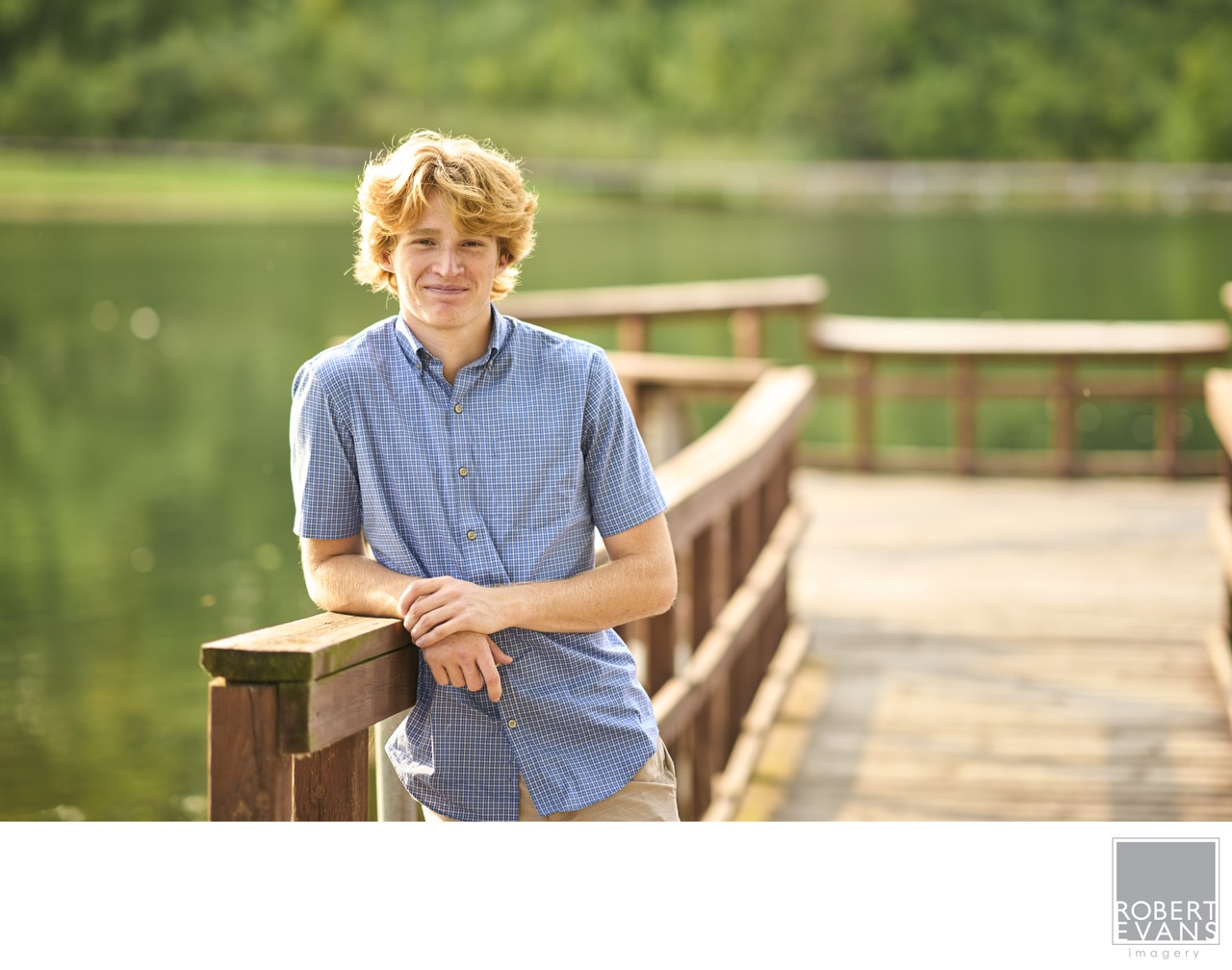 A young man leans against a wooden dock railing beside calm water.
