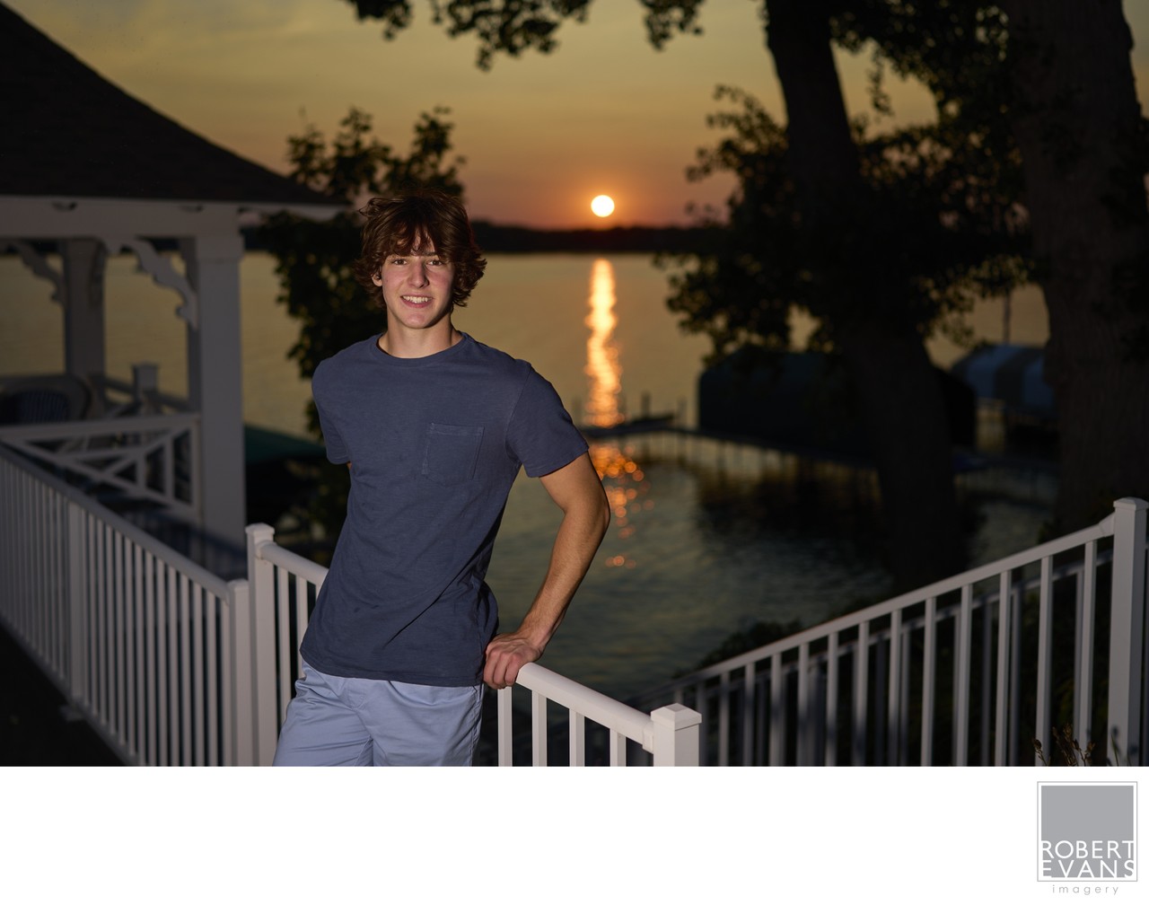 A young man stands beside a waterfront home with the setting sun behind him.
