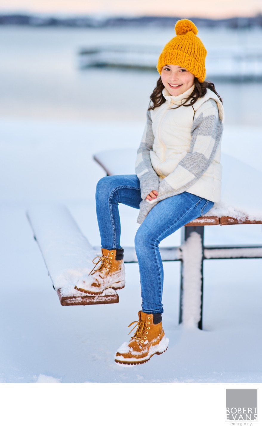 A young woman in a yellow knit hat sits on a snow-covered dock.