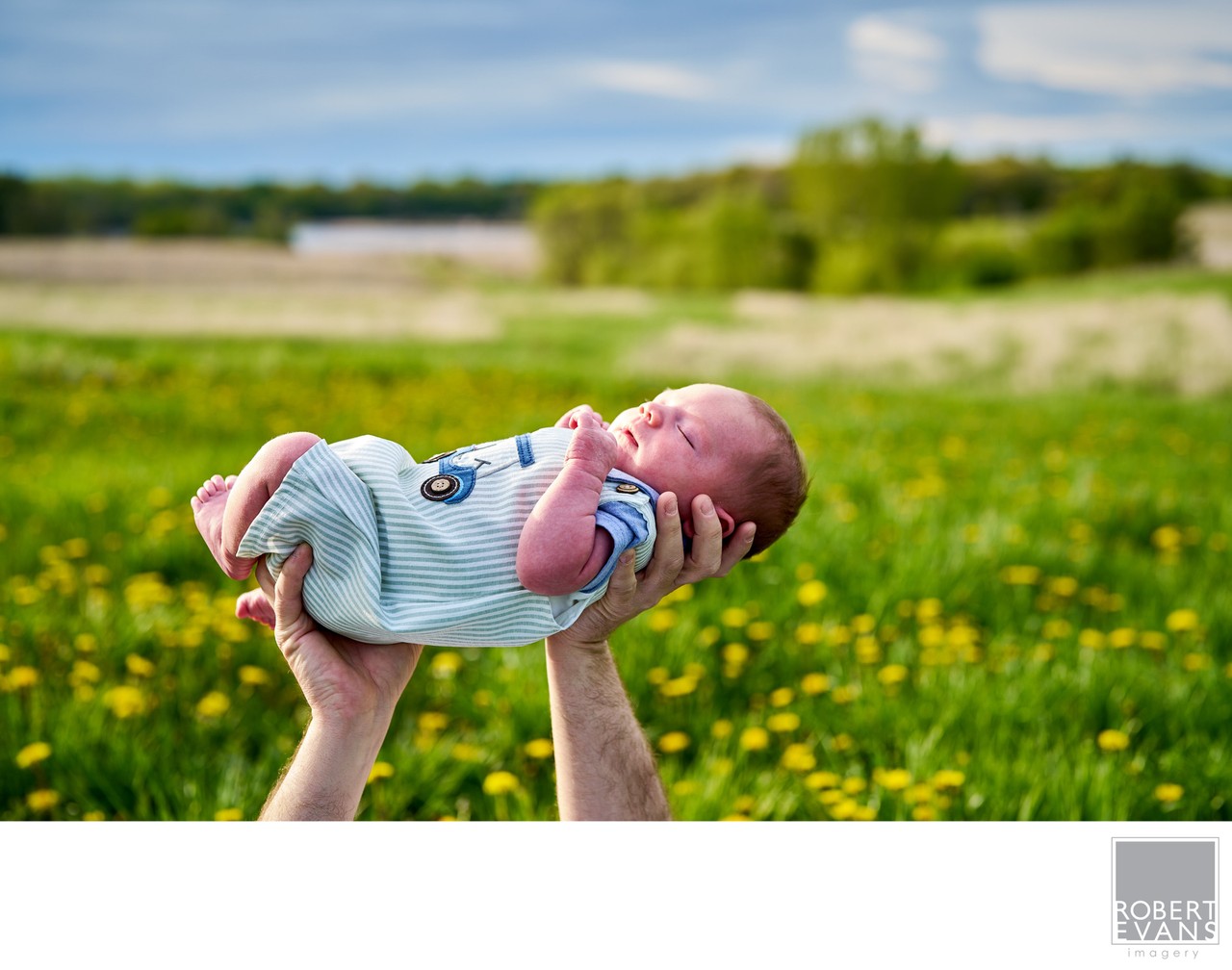 A newborn rests in outstretched hands against a meadow of yellow flowers.