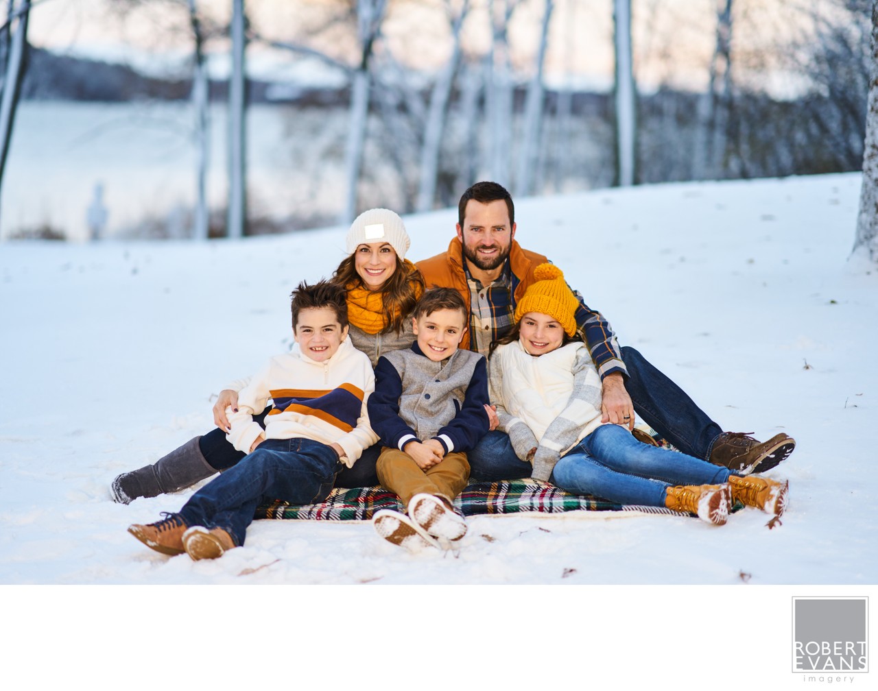 Parents and three children sit close together on a blanket in a snowy woodland setting.
