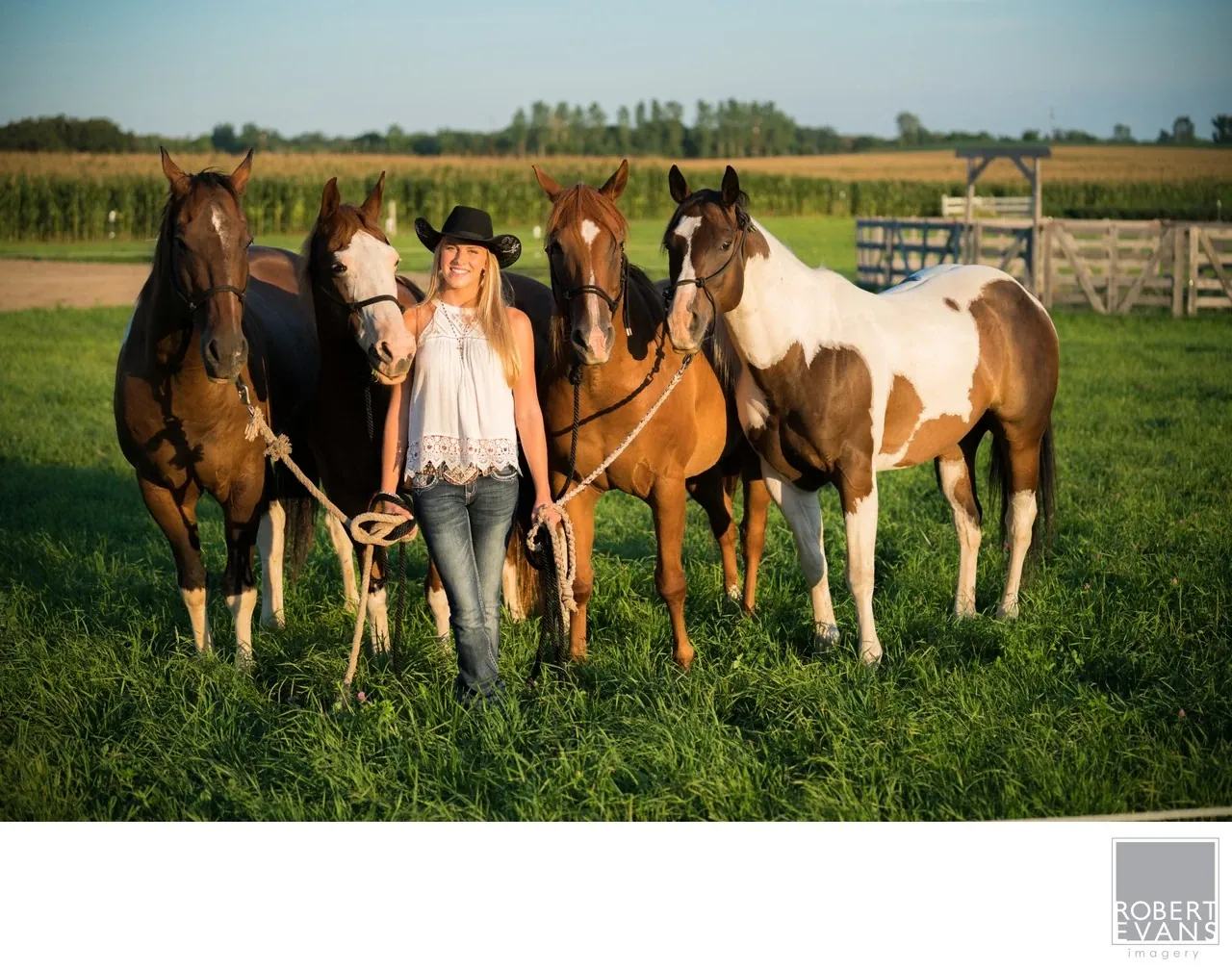 A woman in a cowboy hat stands with four horses in a sunlit pasture.