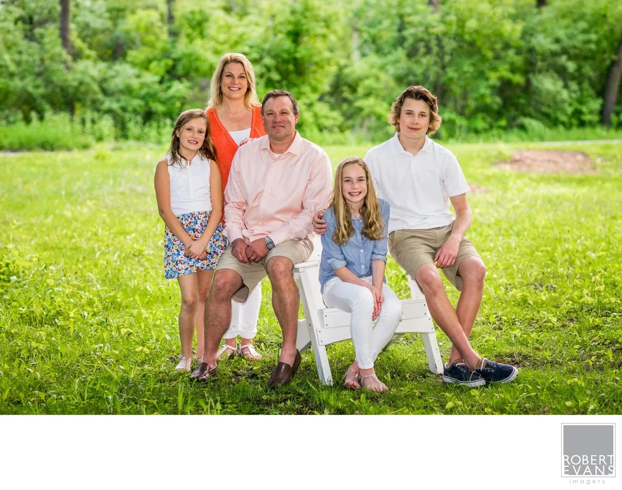 Five family members gather around a seated man on a lawn backed by leafy trees.