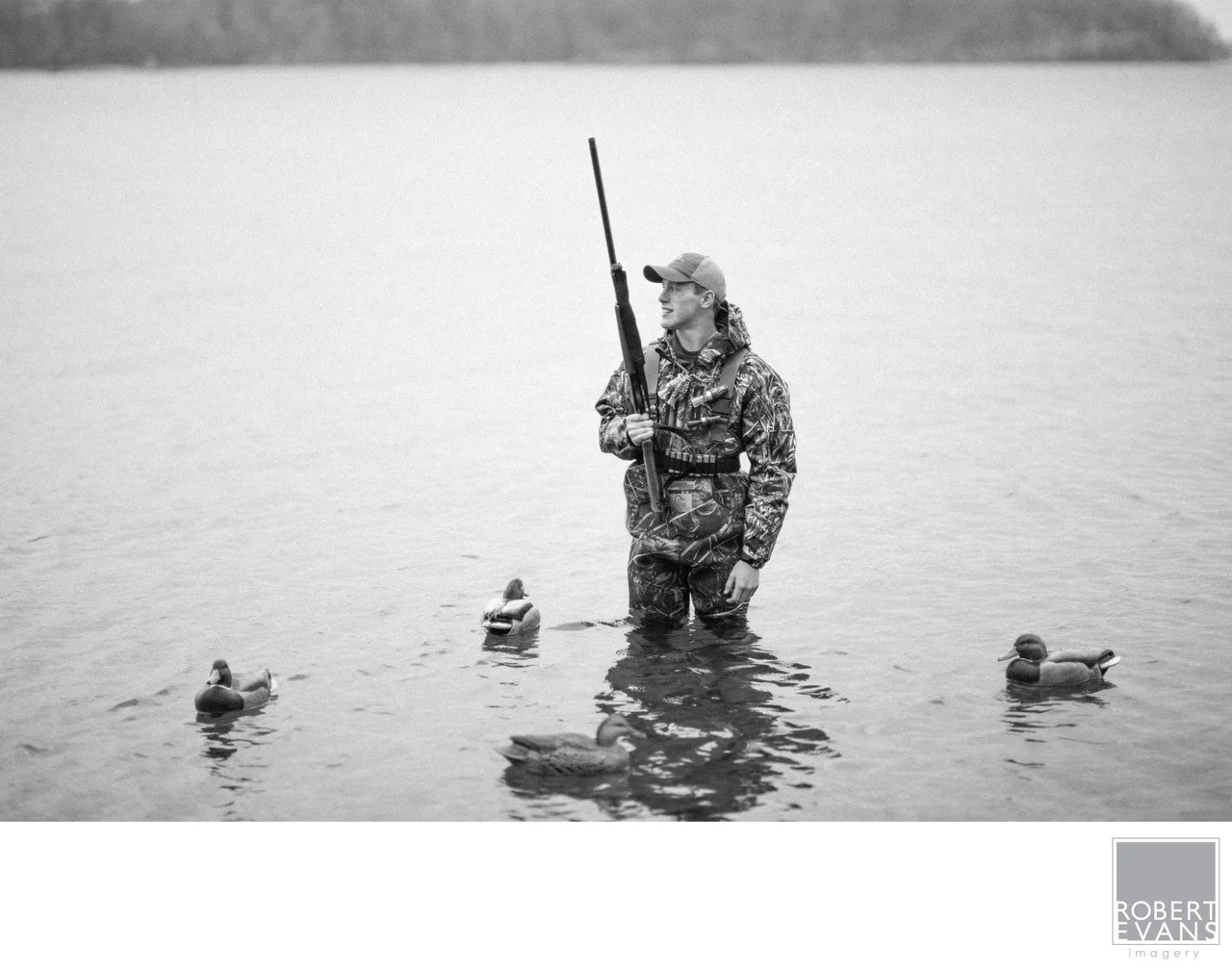A hunter in camouflage stands in shallow water among duck decoys in black and white.