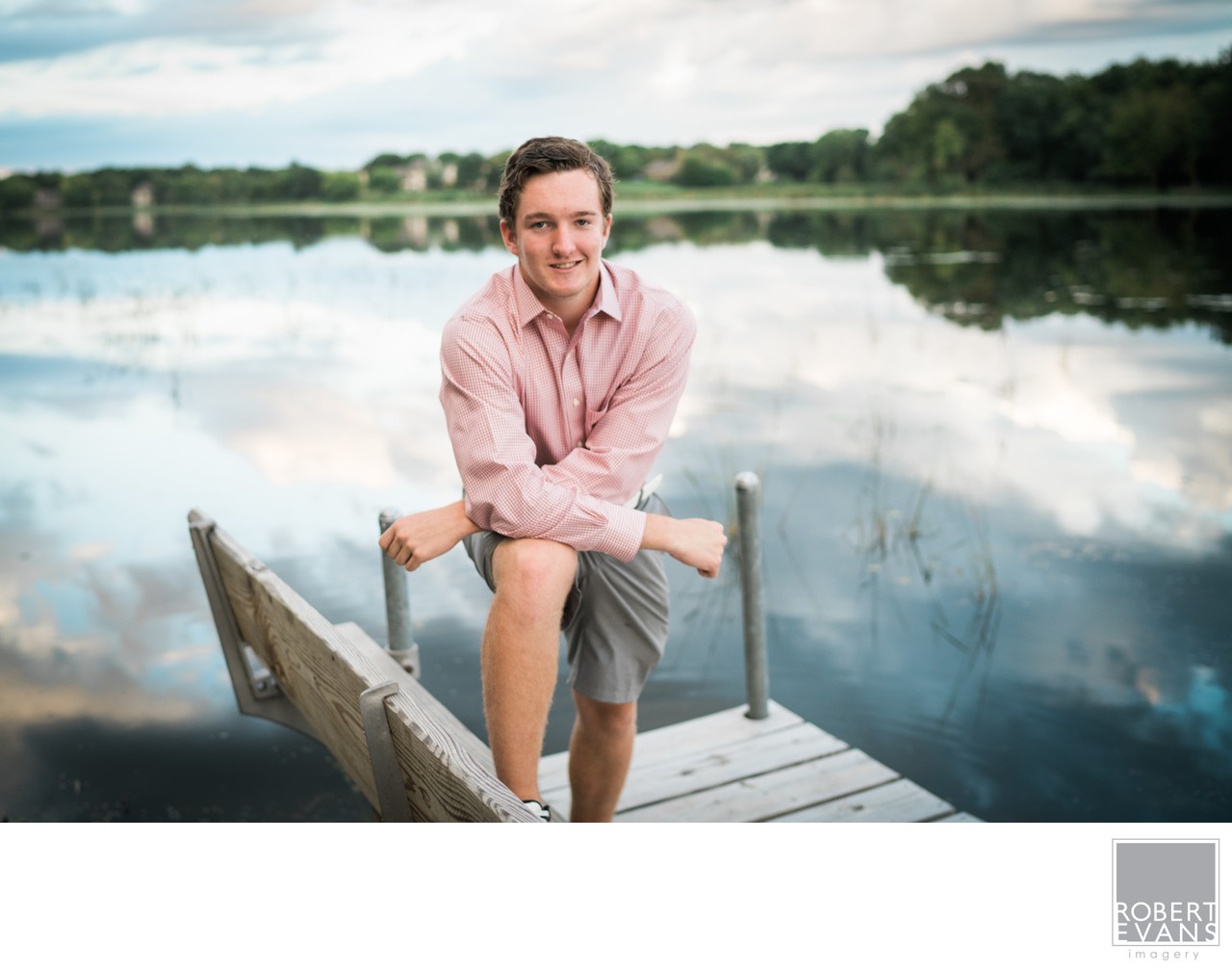 A young man in a pink shirt sits on a wooden dock above still water and reflections.