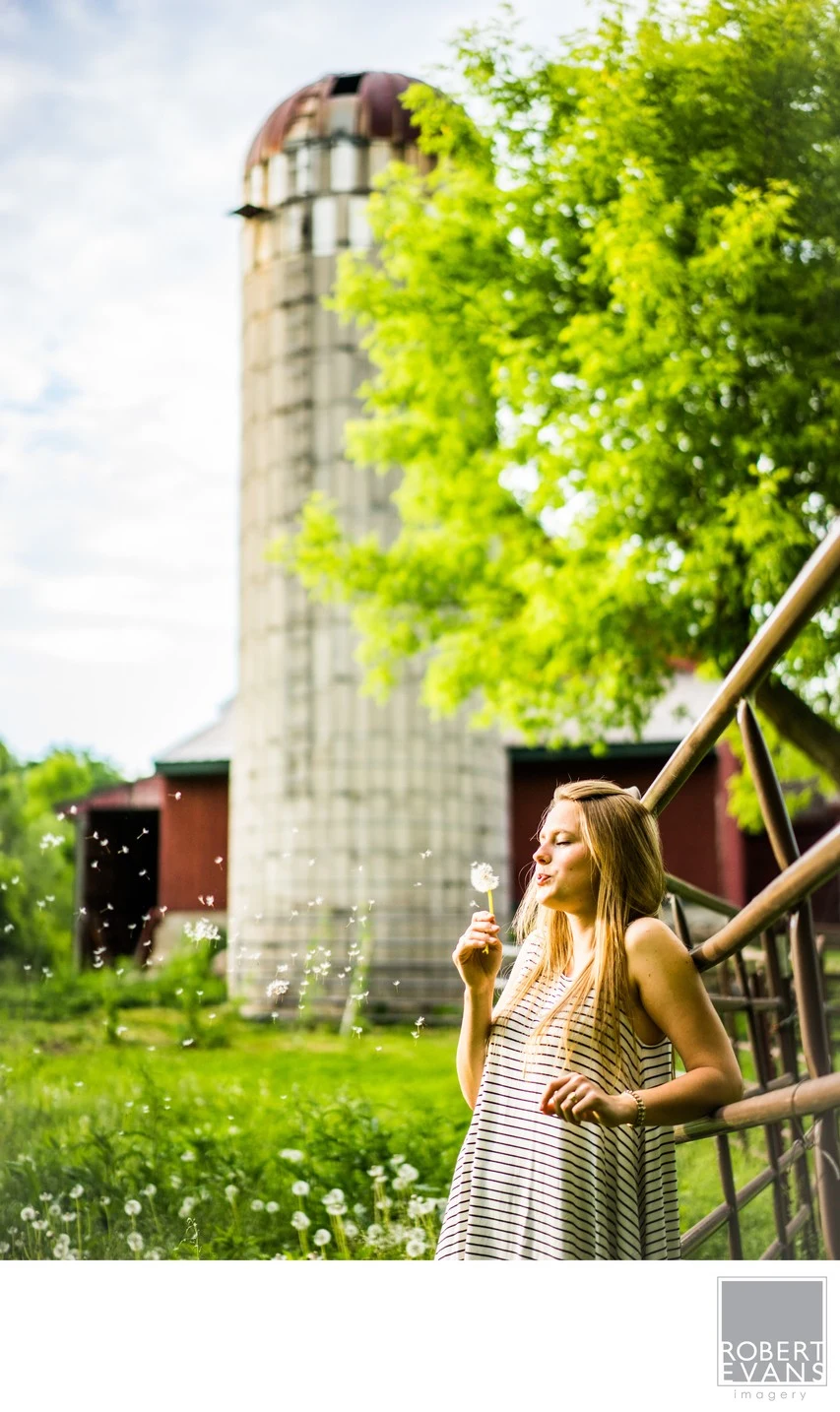 A young woman stands beside a rustic railing with a tall silo and green trees behind her.