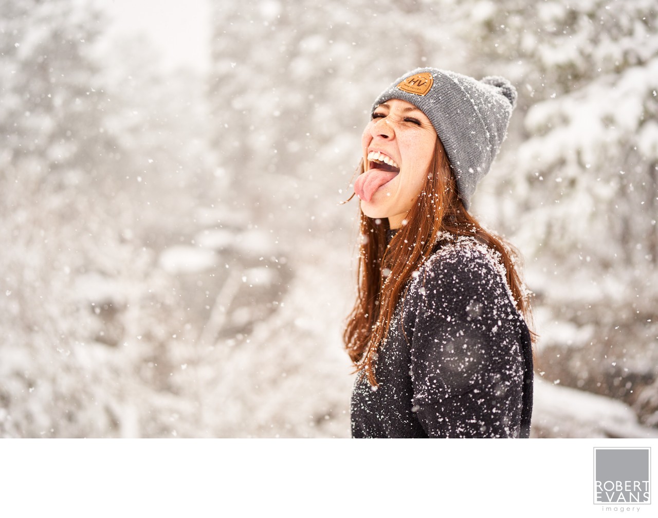 A young woman wearing a knit cap laughs with her tongue out beneath falling snow.
