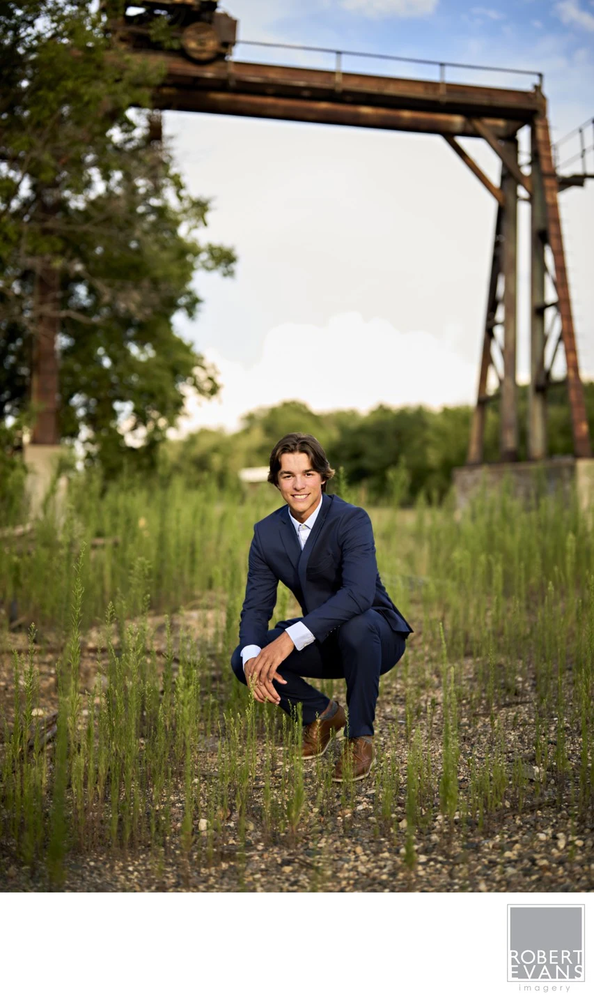 A young man in a dark jacket crouches in tall grass beneath a weathered wooden frame.