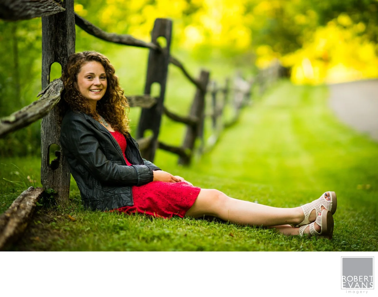 A young woman in a red dress and dark jacket sits on grass beside a wooden fence.