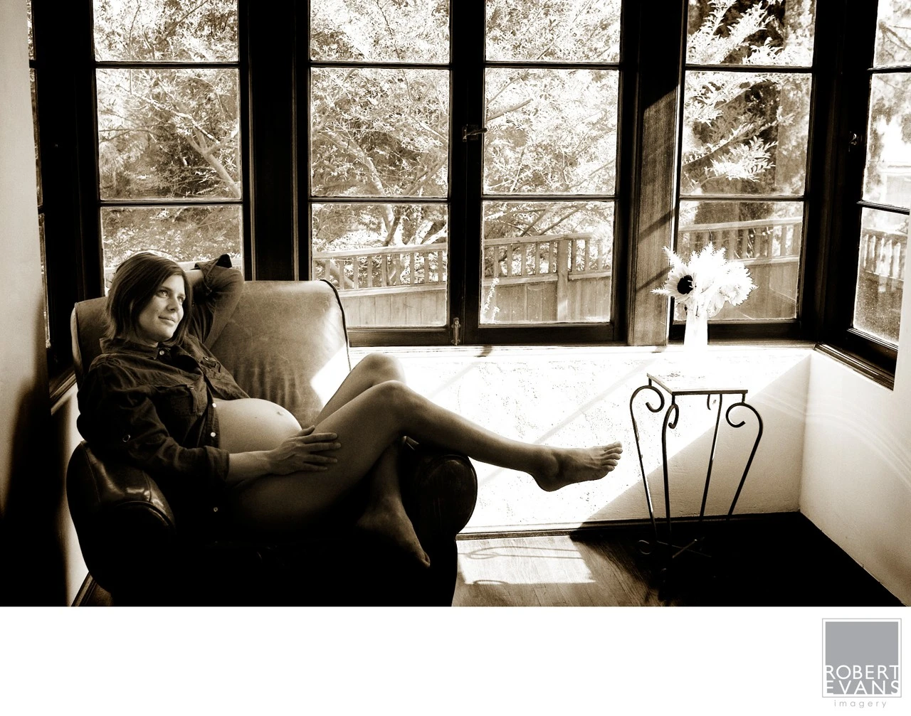 An expectant mother reclines in an armchair beside large windows in a monochrome portrait.
