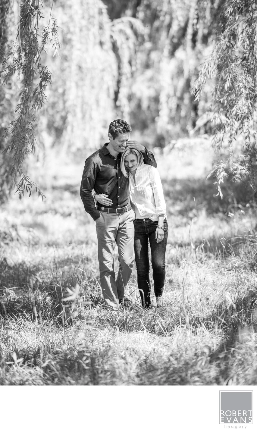 A couple walks close together along a sunlit wooded path in black and white.