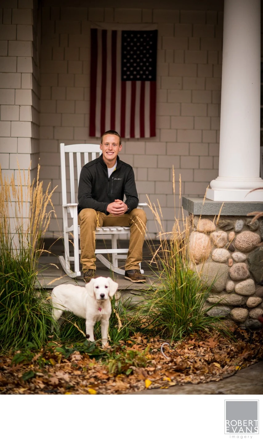 A man sits on a porch beneath an American flag with a white dog in front of him.
