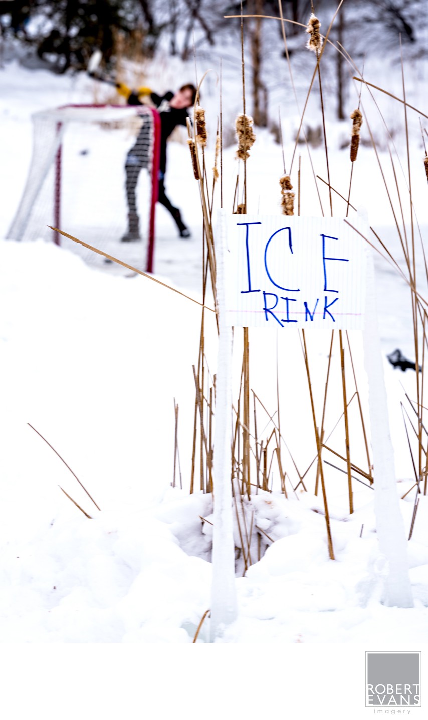 A handmade ice rink sign stands beside a snowy path with a bundled figure in the distance.