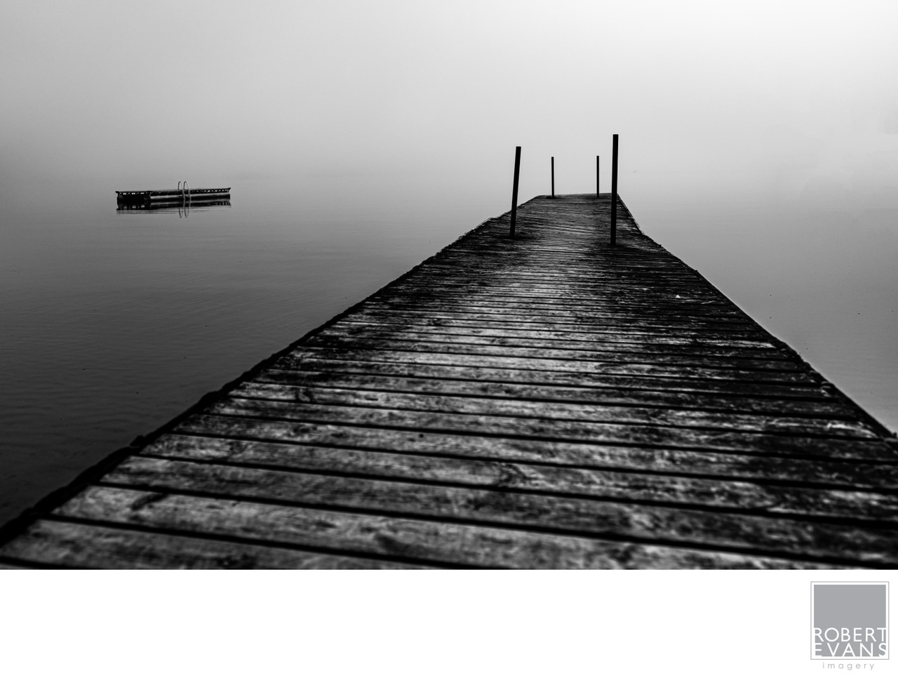 A wooden dock stretches into fog over still water in a minimalist black-and-white photograph.
