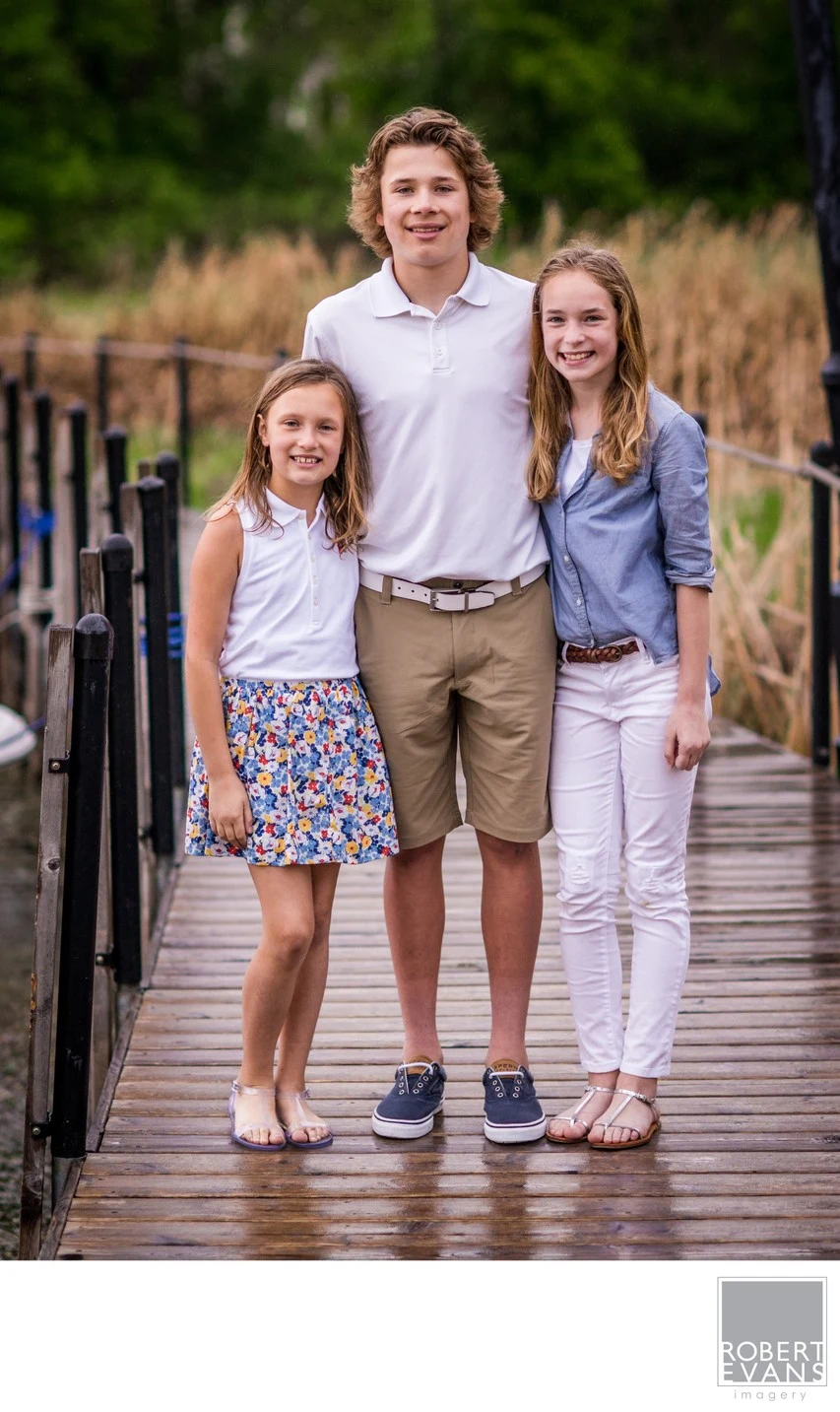 A boy and two girls stand together on a wooden footbridge surrounded by greenery.
