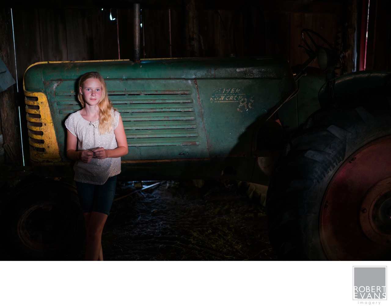 A girl stands beside a large green tractor inside a shaded barn.