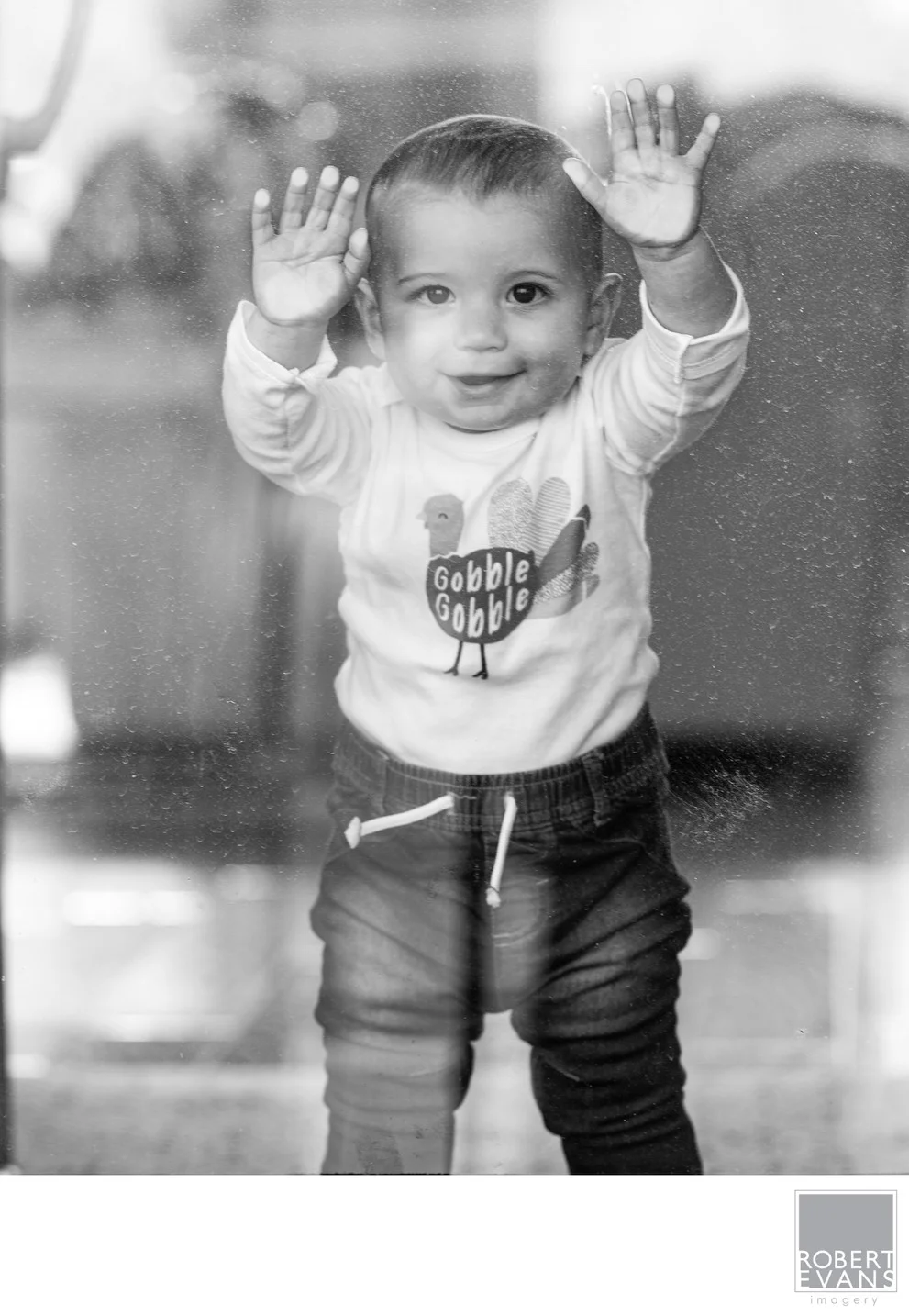 A smiling toddler raises both hands toward the camera in a black-and-white portrait.