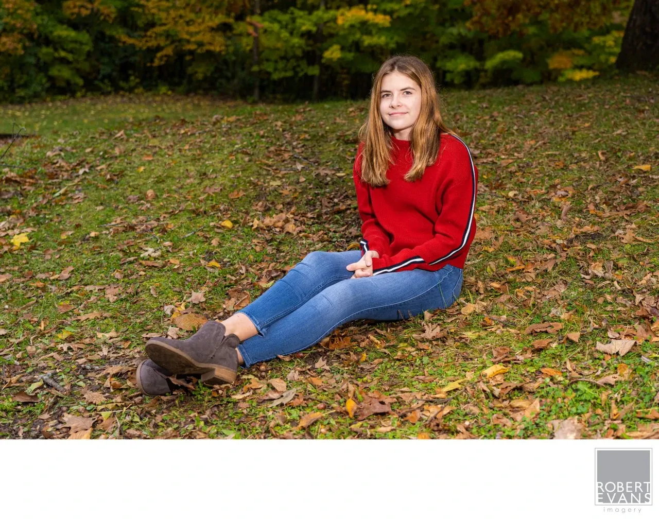 A young woman in a red sweater sits on grass scattered with autumn leaves.