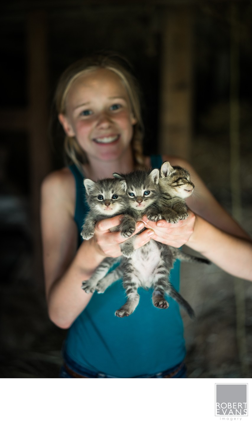 A smiling girl holds two tabby kittens close against a shaded background.