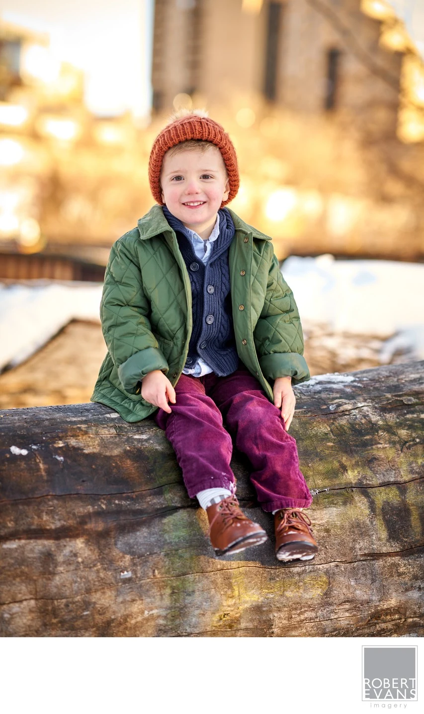 A small child in a green coat and red knit hat sits outdoors in warm light.