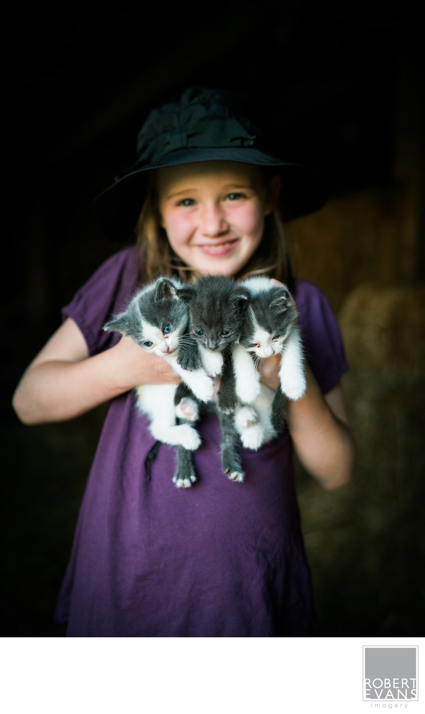 A smiling girl in a dark hat holds two kittens inside a rustic barn.