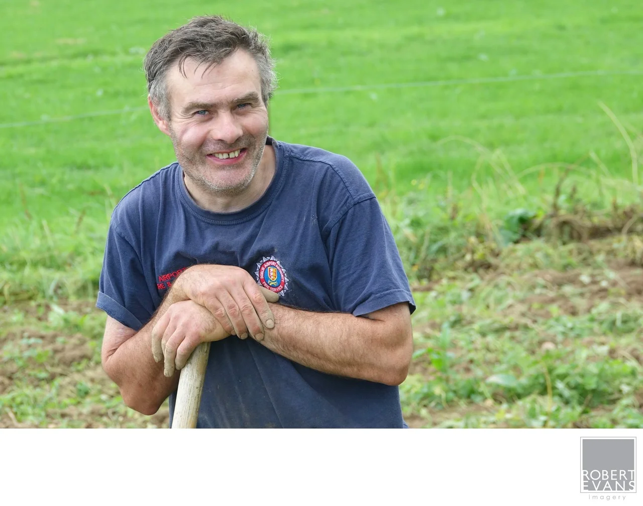 A smiling man in a blue shirt leans on a wooden handle beside a green field.