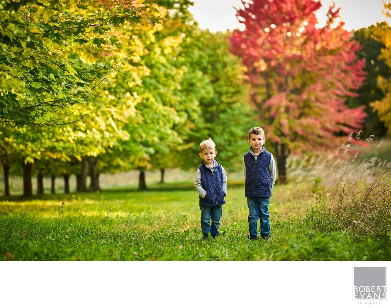 Two boys stand together on a grassy path surrounded by vivid autumn foliage.