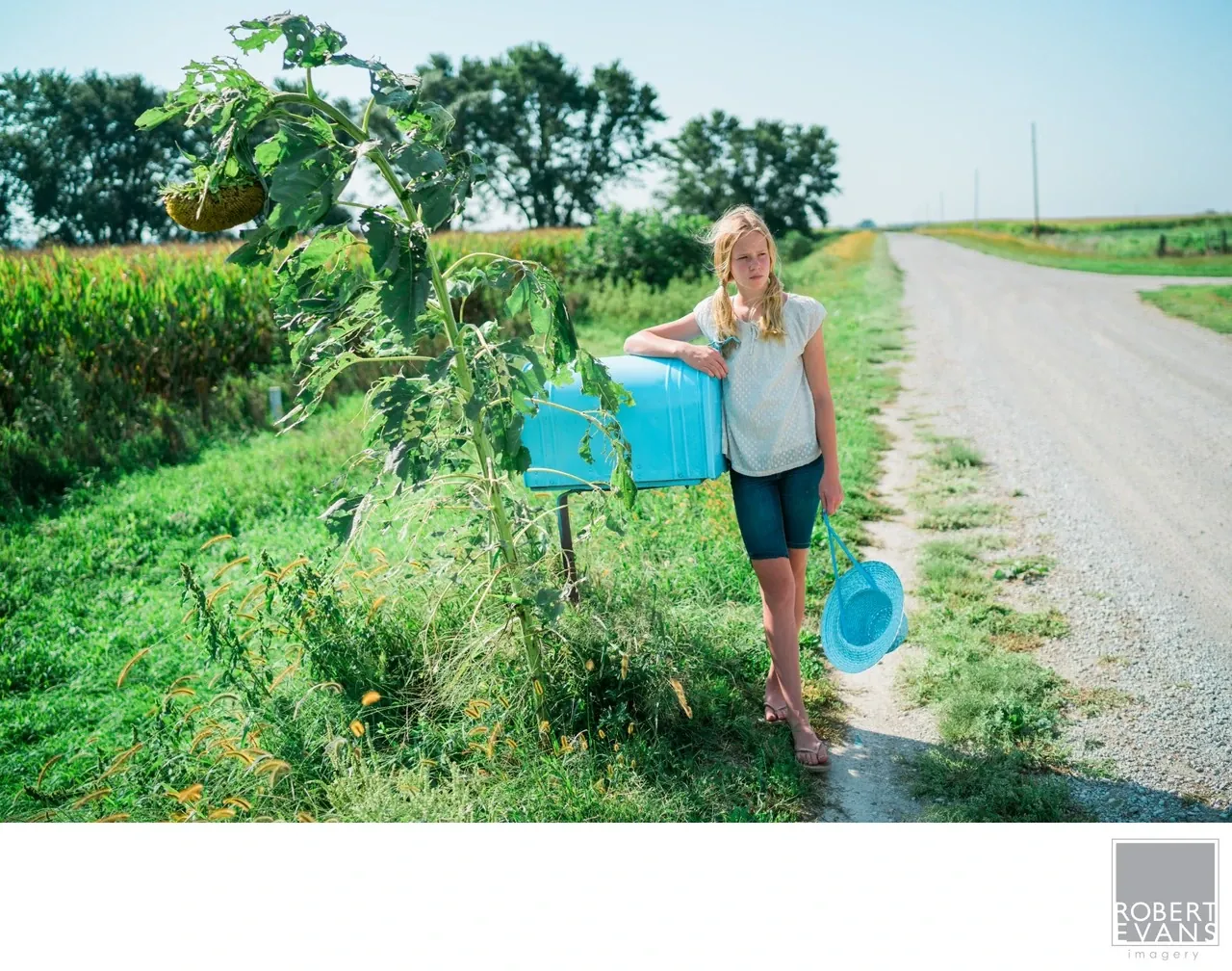 A young woman stands beside a rural road holding a blue container, with fields behind her.