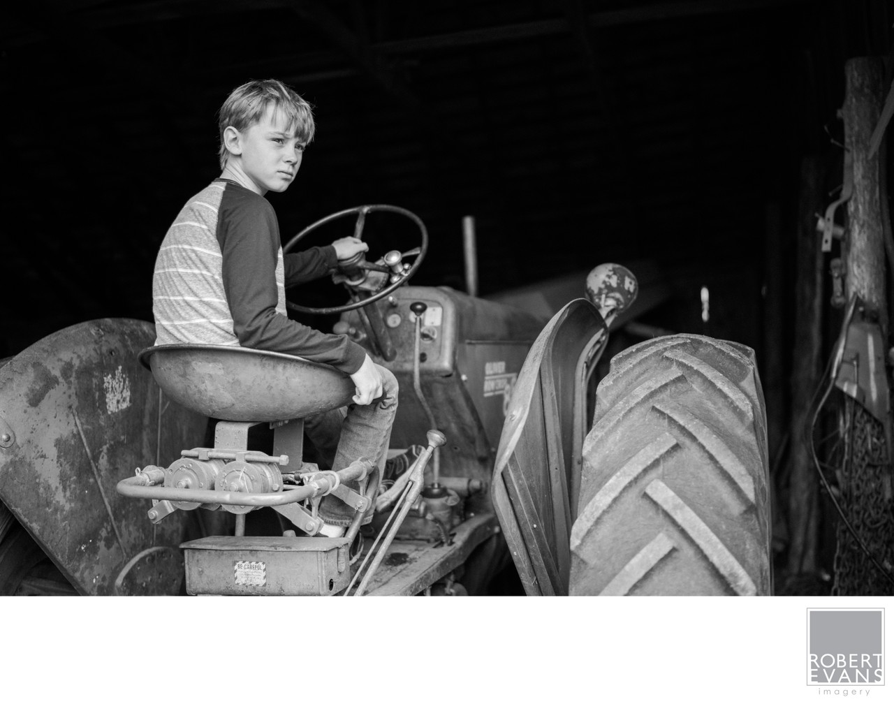 A boy sits at the steering wheel of a tractor inside a barn in black and white.